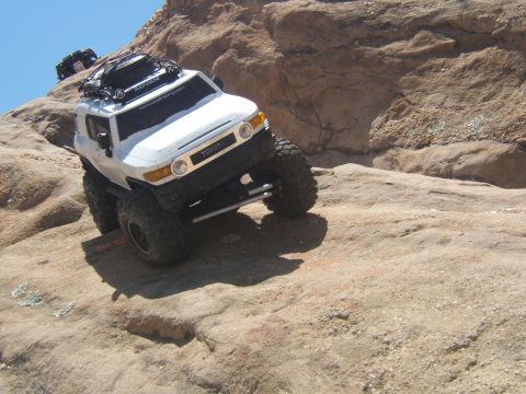 1988 Ford Bronco - A white Toyota FJ Cruiser navigates a rocky incline under a clear blue sky, with another vehicle visible in the background.