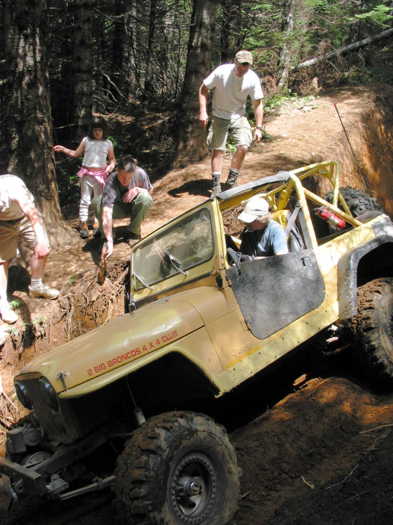 Yellow flat-fender Jeep CJ tilted in a deep rut as several people stand around it in the woods