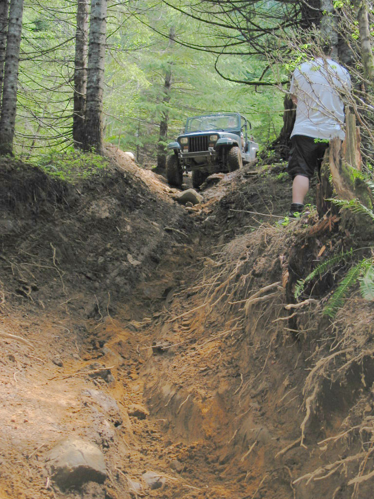 Dark Jeep CJ crawling up a deeply eroded red-dirt trench as a person walks the bank