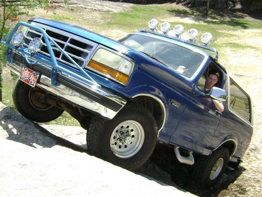 Blue lifted Ford Bronco climbing rocks with roof lights and driver inside