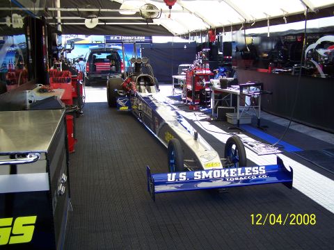 1973 Ford Mustang - A dragster sits in a pit area, ready for action, with a support vehicle parked behind it.