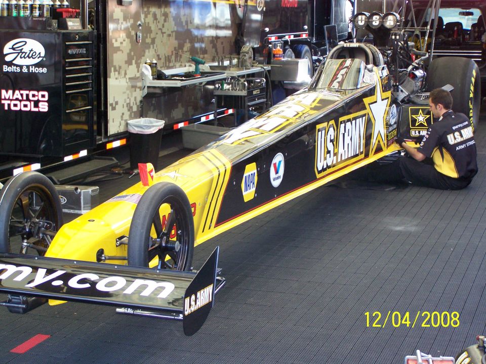 U.S. Army dragster with yellow and black livery in a garage, being worked on by a mechanic