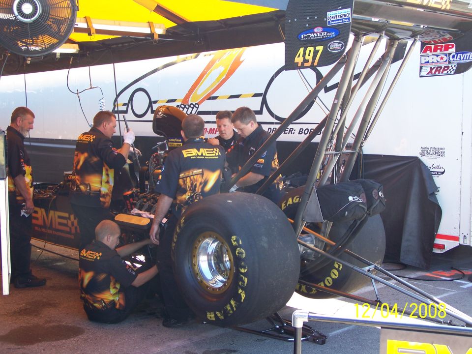 Pit crew works on a dragster, with large rear tires and sponsor decals visible