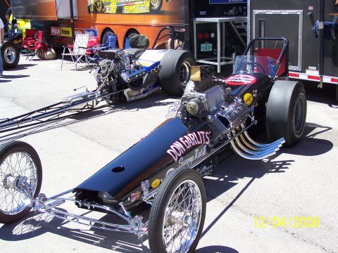1973 Ford Mustang - A black dragster with "DON GARLITS Swamp Rat" lettering is parked next to another dragster, with trailers in the background.