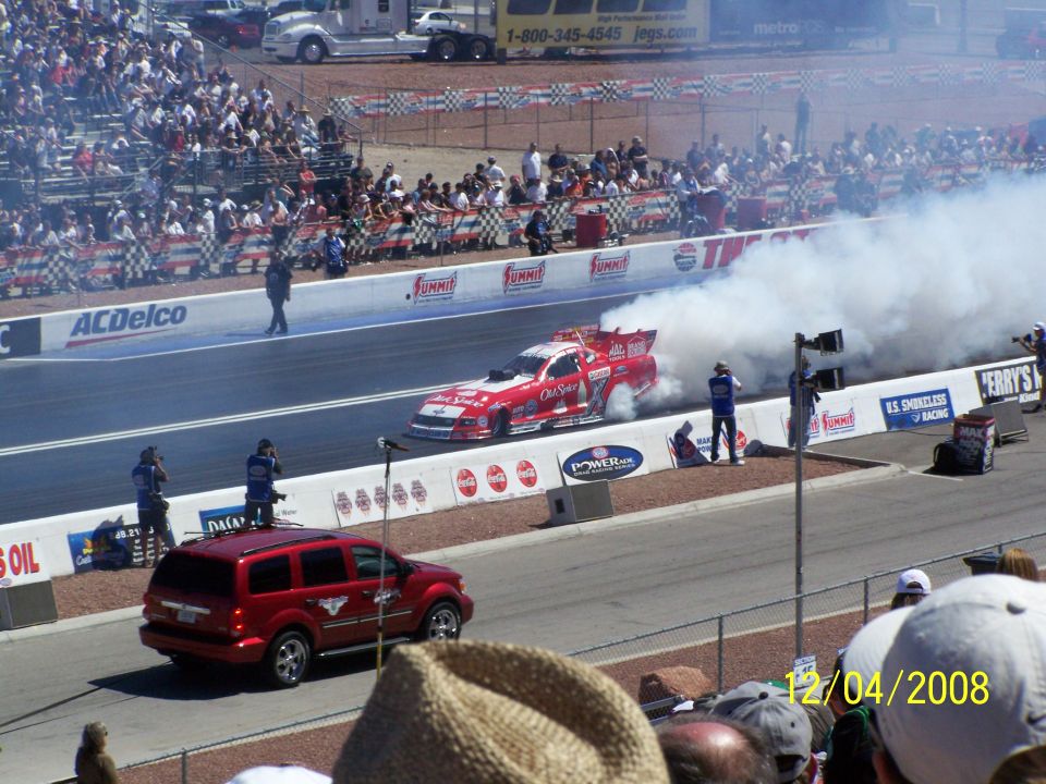 Red drag car doing a burnout on a track, with a red SUV nearby and spectators in the stands