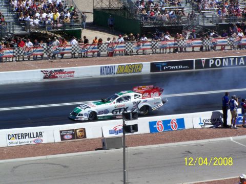 1973 Ford Mustang - A drag racing car competes on a track, with spectators in the stands and sponsor banners lining the course.