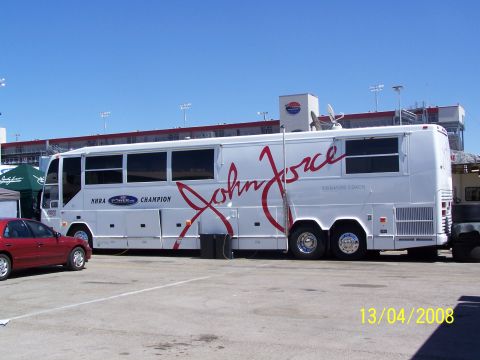 1973 Ford Mustang - A white John Force Signature Coach bus is seen parked next to a red car at a racetrack. The bus displays 'NHRA CHAMPION' and 'John Force SI…