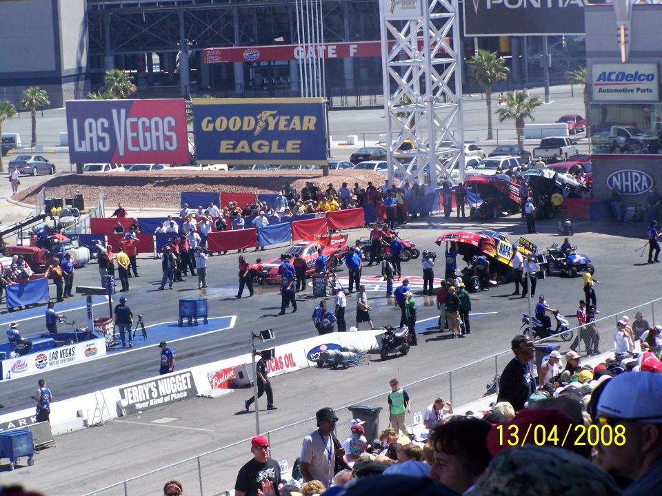 Drag strip scene with various race cars, motorcycles, and spectators under a sunny sky