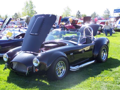 1973 Ford Mustang - A black Cobra-style sports car with its hood open is parked on a grassy field at an outdoor car show. Another blue sports car is visible be…