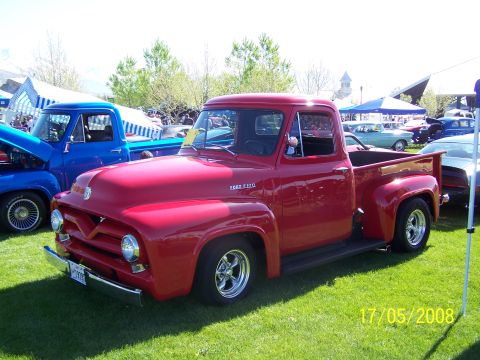 1973 Ford Mustang - A bright red 1950s Ford F-100 pickup truck is parked on a grassy field in front of other vehicles and tents. The truck is shown from a thre…