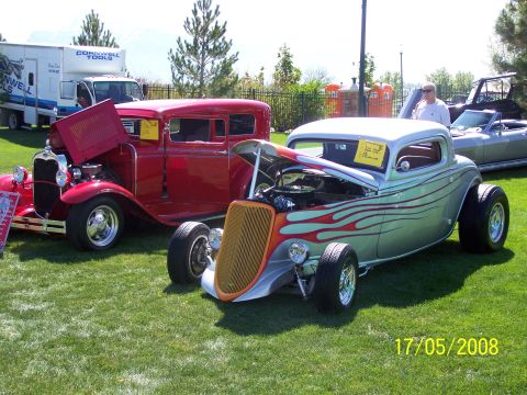 1973 Ford Mustang - Two classic hot rod cars are parked on a grassy lawn. The car in the foreground is silver with red flames and has its hood open, while the…
