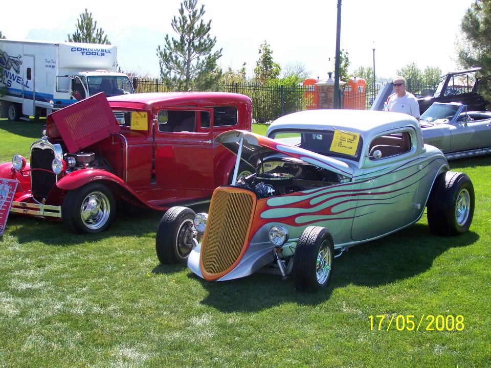 Two classic hot rod cars, one silver with flames and one red, parked on grass with hoods open.