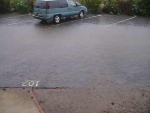 1990 Ford Bronco - A teal minivan is parked in a flooded parking lot during a rainstorm. The parking space number "102" is visible on the wet asphalt.