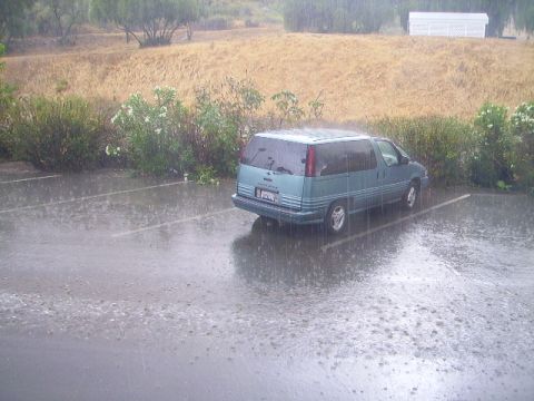 1990 Ford Bronco - A teal minivan is parked in a wet parking lot during a rainstorm. The vehicle is viewed from a rear three-quarter angle.