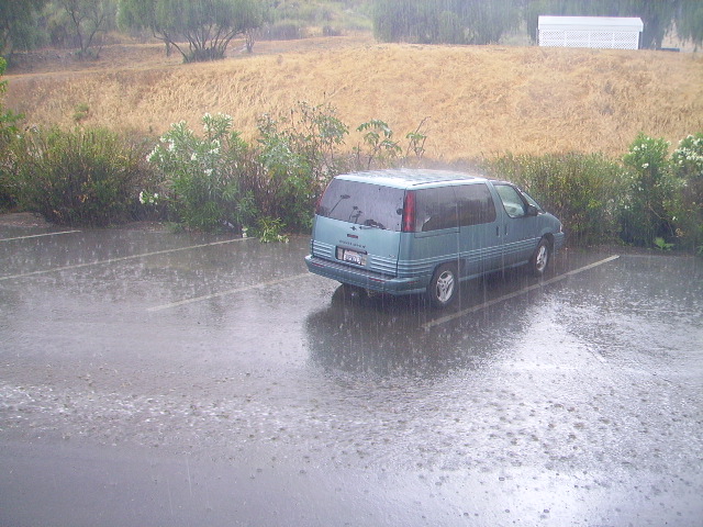 Teal minivan parked in a wet parking lot during a rainstorm, rear three-quarter view.