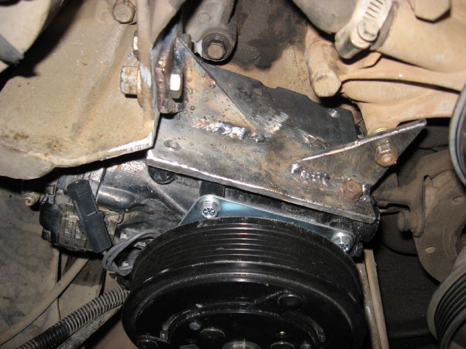 Custom metal bracket holding an AC compressor on a vehicle engine.