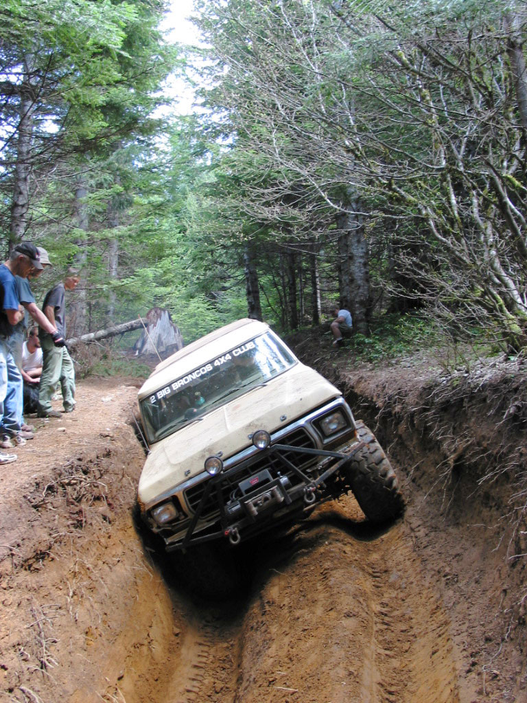 Ford Bronco stuck in a dirt ditch on a wooded trail, with people watching