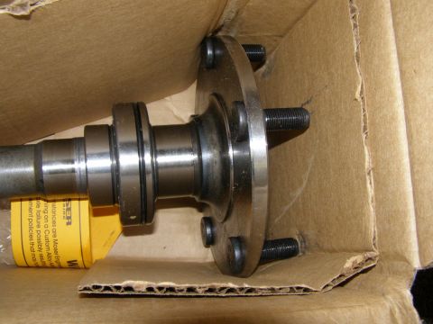 1993 Ford Bronco - A close-up view of a metal axle shaft with a flange and four studs, resting inside a cardboard box. A yellow warning label is partially vis…