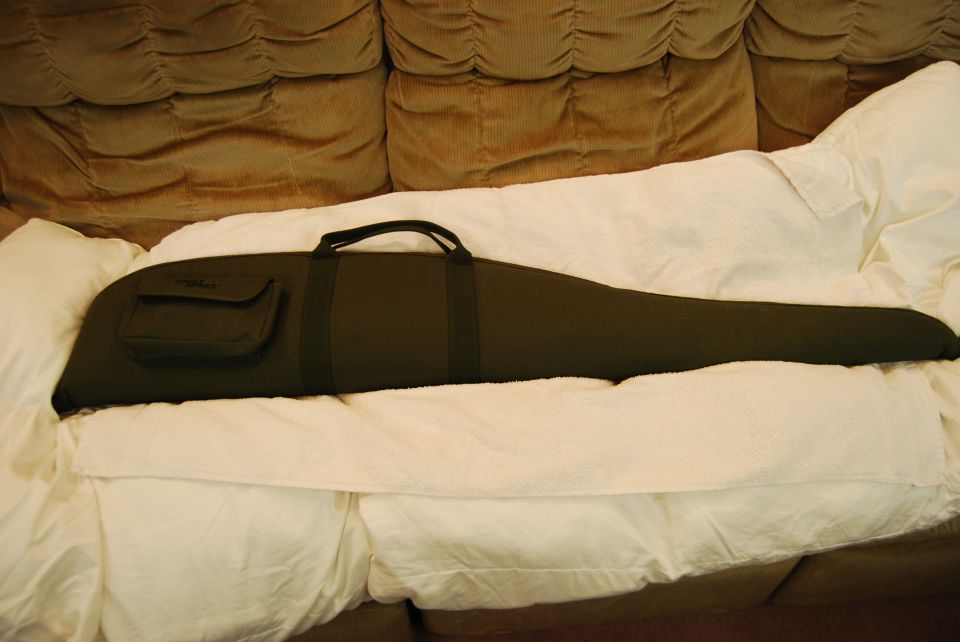 Dark green gun case on white bedding with a brown couch behind.
