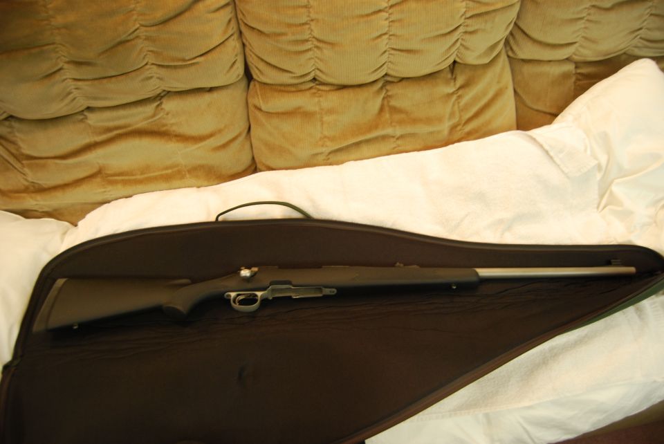 Rifle in a gun case on a white blanket with a couch behind it.