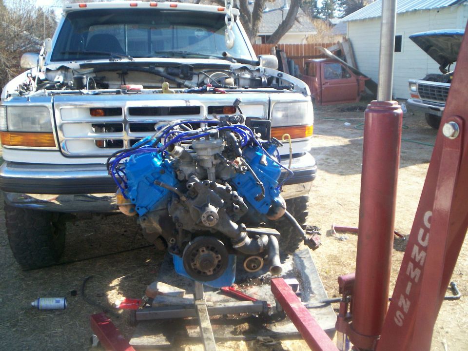V8 engine with blue valve covers on a red hoist in front of a white Ford pickup with empty engine bay