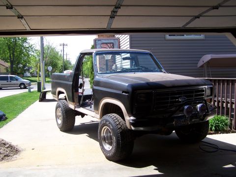 1986 Ford Bronco - A dark-colored Ford Bronco is parked in a garage with the door open. The vehicle is missing its doors and has aftermarket lights mounted on…