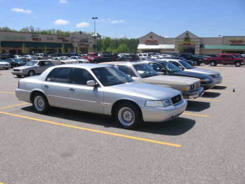 1988 Mercury Grand Marquis - Silver Ford Crown Victoria sedan on lace-spoke wheels parked in a strip-mall lot, front three-quarter view, with a tan Mercury sedan and ot…