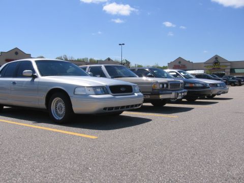 1988 Mercury Grand Marquis - A row of sedans are parked in a lot on a sunny day. The closest vehicle is a silver sedan with a chrome grille and wire-spoke wheels.