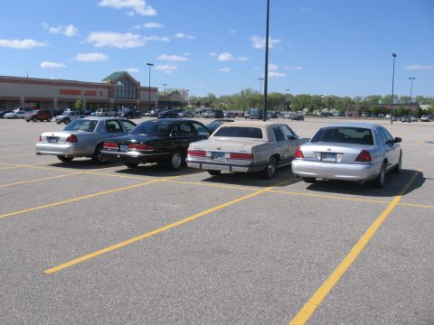 1988 Mercury Grand Marquis - Four sedans are parked in a paved lot in front of a strip mall. The vehicles are a silver sedan, a black sedan, a tan sedan, and a silver s…