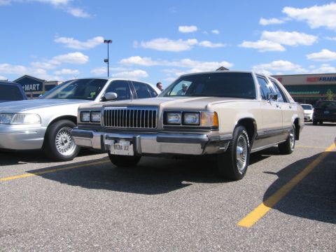1988 Mercury Grand Marquis - A tan 1988 Mercury Grand Marquis is parked in a lot under a blue sky with scattered clouds. The car features a chrome grille and a license…