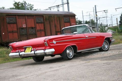 1991 Ford Crown Victoria - A vintage red convertible with prominent tail fins is parked on a gravel surface near a rusty train car.