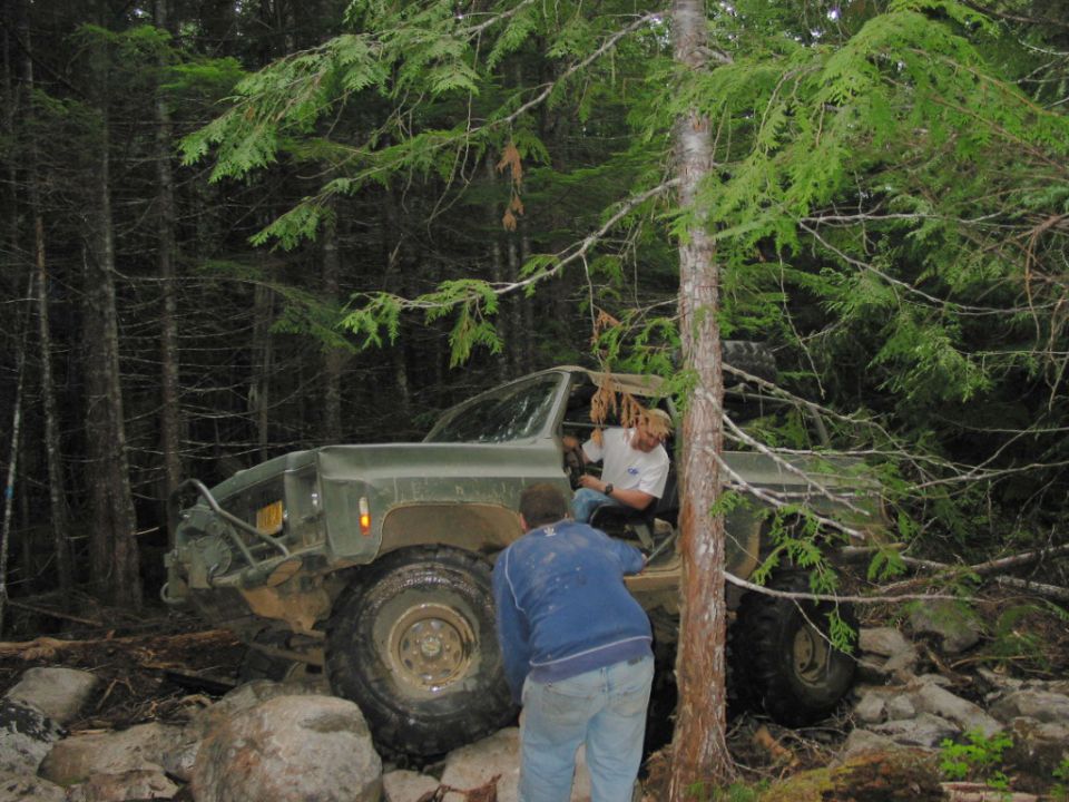 Olive green off-road vehicle with large tires, two people visible, in a wooded area