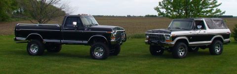 1979 Ford Bronco - Two lifted trucks, a black pickup and a black and white Bronco, sit on a grassy field with a harvested field in the background.