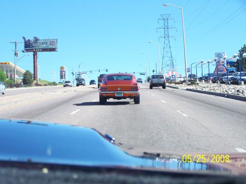 1972 Ford Ranchero - Orange 1969 Ford Mustang coupe seen from behind on a sunny day, with other vehicles and a car dealership in the background.