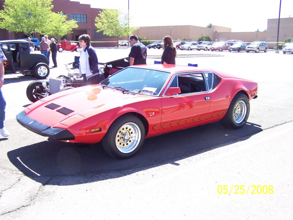 Red sports car with black front bumper and gold-colored wheels. Other cars and people are visible in the background
