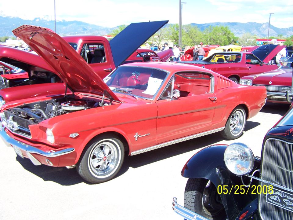 Red 1965 Ford Mustang coupe with hood open, parked at a car show