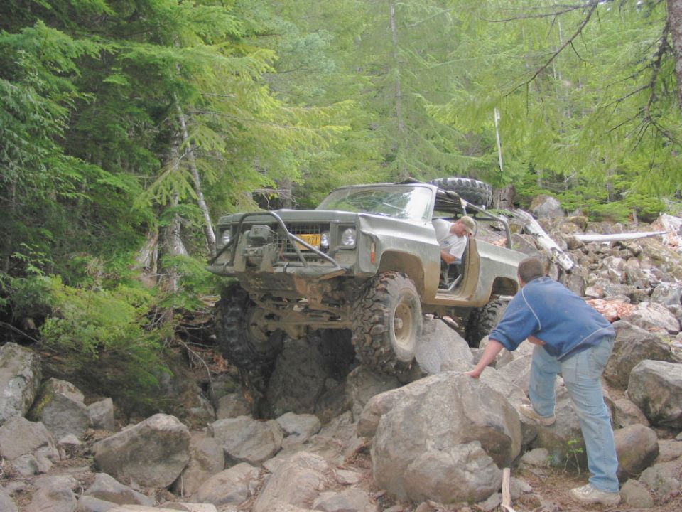 Off-road truck with large tires navigating a rocky trail. A person is climbing out of the driver's side