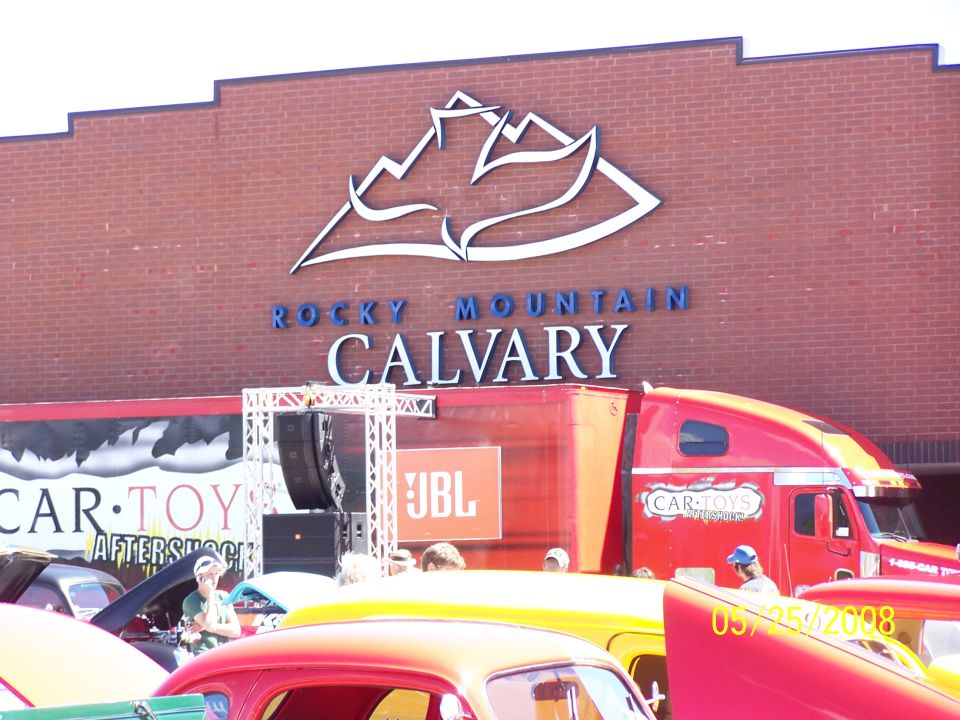 Red semi-truck with 'CAR-TOYS' logo parked at a car show in front of a brick building with 'ROCKY MOUNTAIN CALVARY' sign. Several classic cars are visible