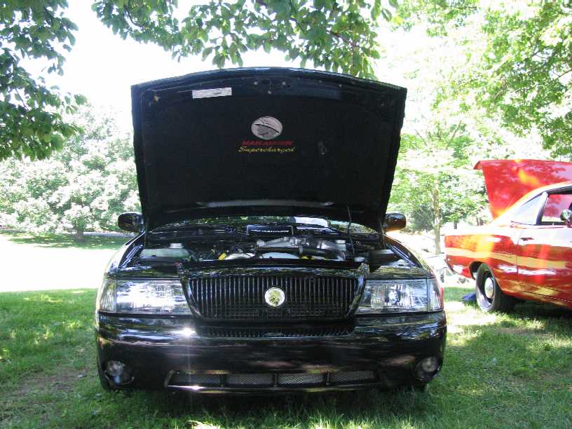 Front view of a black Mercury Marauder with hood open, parked on grass with a red car to the side.