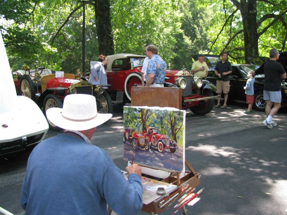 Artist painting a vintage red car at an outdoor car show with other cars in the background.