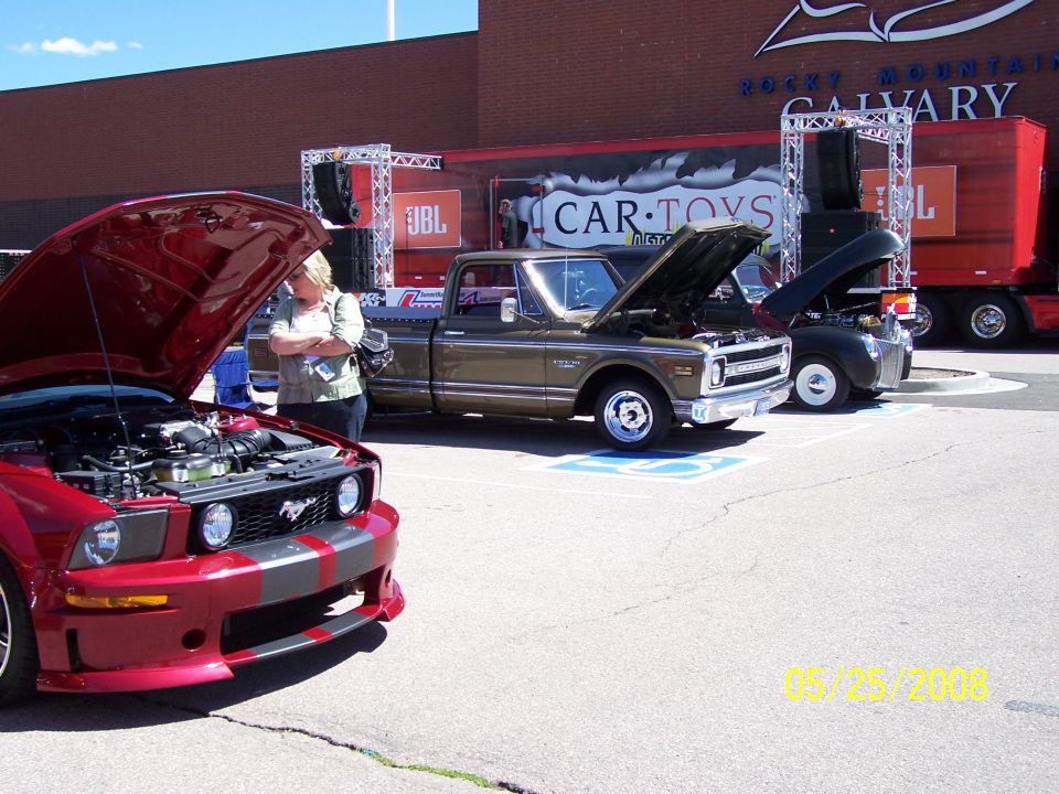 Red Ford Mustang with hood open, parked next to a brown pickup truck. Woman looking at engine.