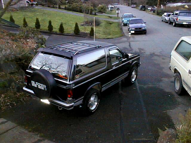 Black two-door Blazer SUV, rear 3/4 view, with spare tire cover and roof rack in a driveway