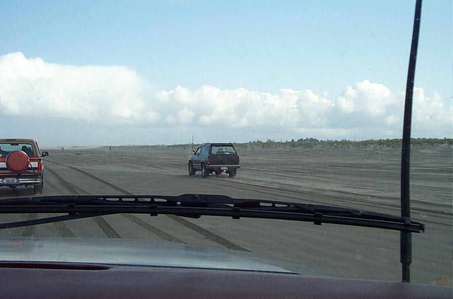 Dark SUV driving on a sandy beach seen through a windshield, with a parked pickup at left.
