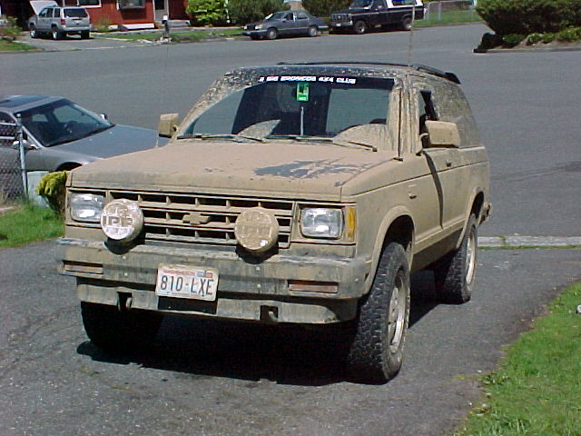 Muddy tan two-door Chevy SUV with off-road tires and front auxiliary lights, 3/4 front view