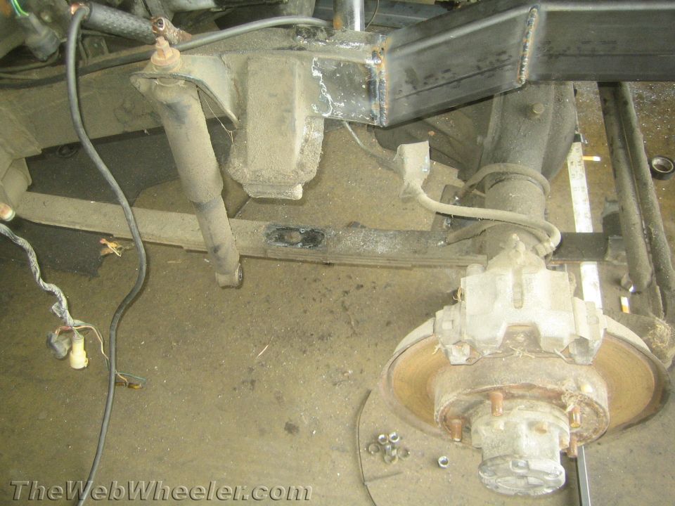 Vehicle chassis with leaf springs, shock absorber, and rear axle assembly. Rusty brake rotor visible