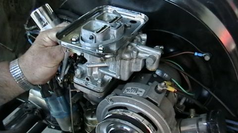1970 Volkswagen Beetle - A person works on the carburetor of a Volkswagen Beetle engine, with a visible metal tag on the engine block.