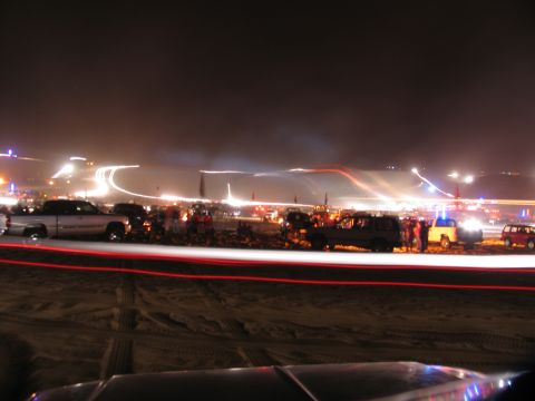 1984 Ford Bronco - A nighttime scene captures multiple vehicles gathered on sandy terrain, illuminated by distant lights and vehicle headlights.