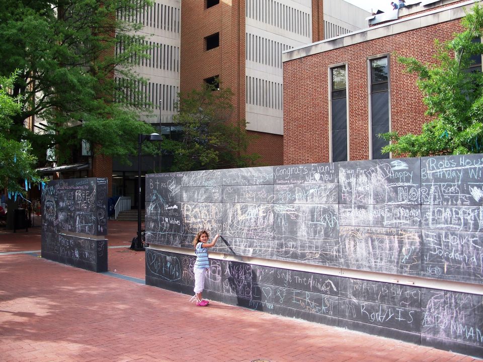 Child writing on a public chalkboard wall covered in messages on a brick plaza