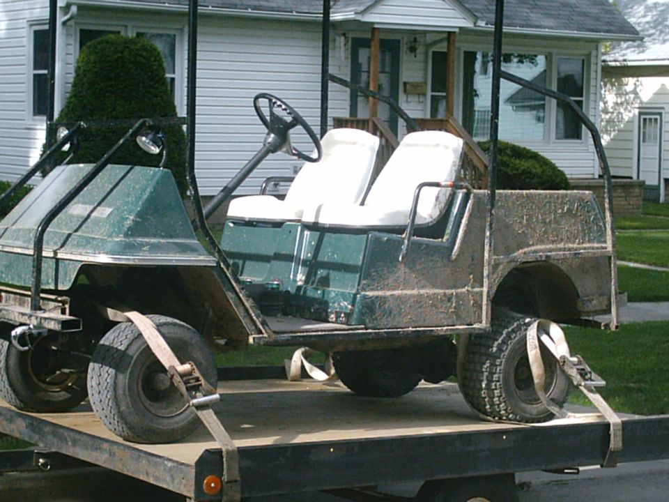 Green golf cart with white seats strapped on a flatbed trailer in a residential yard