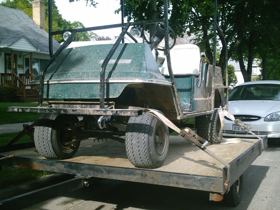 Green golf cart with black frame strapped on a flatbed trailer in a residential street